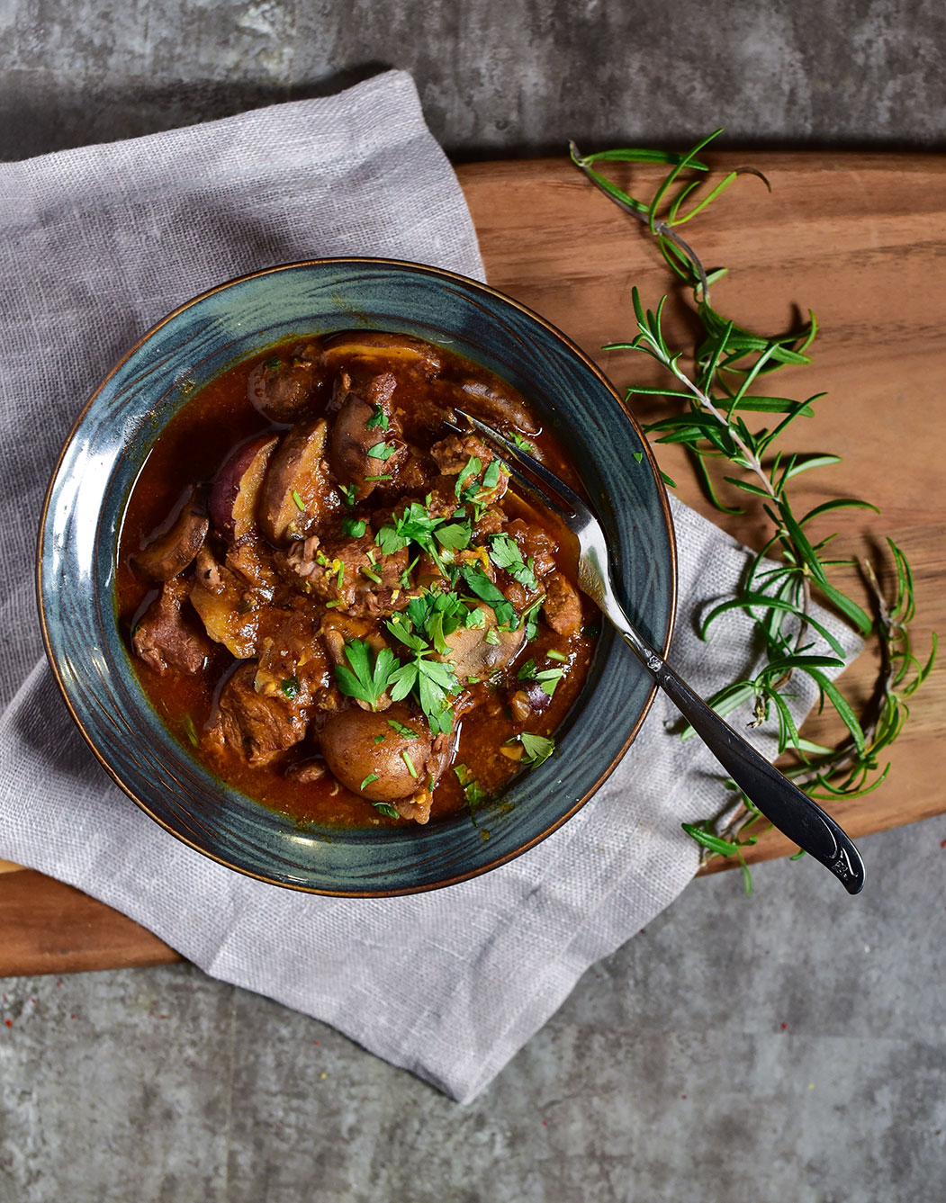 top view of a bowl of beef stew on a wooden cutting board with rosemary and a fork