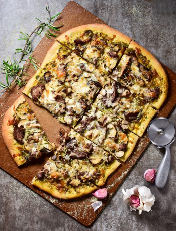 image of a pizza with a pesto sauce and mushrooms with rosemary on the side, garlic cloves and a pizza cutter